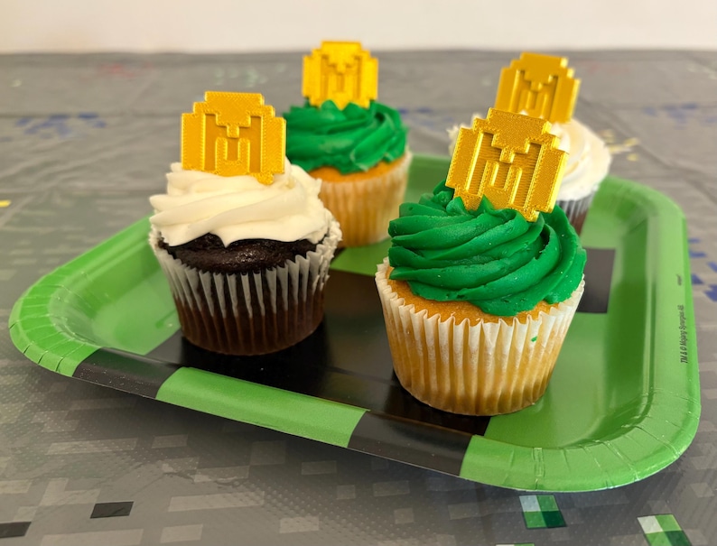 May include: Four cupcakes decorated with green frosting and gold Minecraft-themed toppers on a green and black Minecraft-themed paper plate.