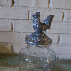 Pode incluir: Um frasco de vidro transparente com tampa prateada com uma figura detalhada de galo. O frasco tem detalhes verticais canelados e uma base arredondada. O galo está empoleirado na tampa, adicionando um toque decorativo ao recipiente.
