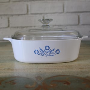 Puede incluir: Cazuela blanca con tapa de cristal transparente. El plato presenta un diseño floral azul. El plato está sobre una superficie decorativa frente a una pared de ladrillos blancos.