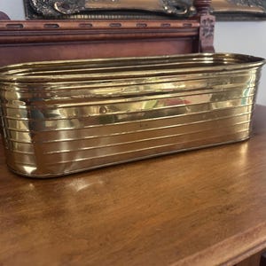 May include: An elongated, oval-shaped brass planter featuring a series of horizontal, embossed ridges along its exterior walls. The metallic surface has a polished, reflective finish with a warm golden tone and a smooth, rounded top rim.