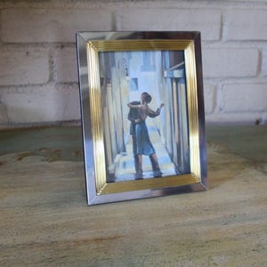 May include: A rectangular picture frame with a silver and gold border. The frame contains a painting of a couple dancing in a narrow street. The painting has a blue and white colour scheme.