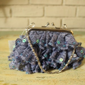 May include: A small, ruffled, lavender-colored purse with a silver clasp and a delicate chain strap. The purse is adorned with sequins in shades of blue and green, adding a touch of sparkle. The purse is approximately 6 inches wide.
