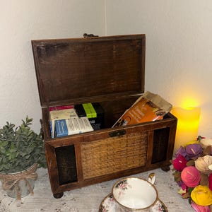 May include: A wooden tea chest with a hinged lid, containing tea boxes and bags. The chest has a woven front panel and sits on small feet. A teacup and saucer with floral designs are in front of the chest.
