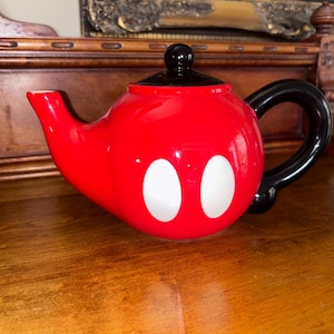 Può includere: Una teiera in ceramica rosso brillante con un coperchio nero lucido dotato di pomello rotondo e un manico nero curvo. Il corpo della teiera è decorato con due grandi forme ovali bianche verticali sulla parte anteriore, che ricordano il design di un personaggio.