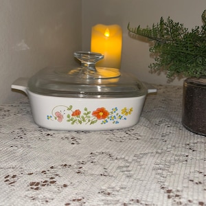 May include: White casserole dish with a clear glass lid. The dish features a floral design with orange, yellow, and blue flowers. The dish is on a white lace tablecloth. A lit candle and a small plant are in the background.