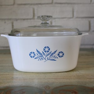May include: White casserole dish with a clear glass lid. The dish features a blue floral design. The handles are on either side of the dish. The lid has a clear glass knob. The dish is sitting on a wooden surface.