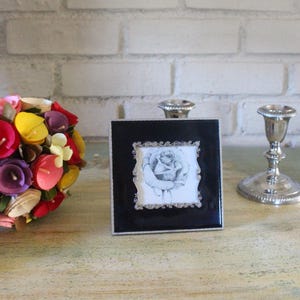 May include: A colorful bouquet of paper flowers with red, yellow, pink, and purple petals. A black framed picture with a rose drawing and two silver candlesticks are also present.