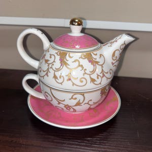 Puede incluir: Un juego de té de porcelana blanca para una persona, que incluye una tetera apilada sobre una taza y un plato. El conjunto está decorado con un patrón de filigrana dorada y detalles en rosa vibrante en la tapa, la base de la taza y el borde del plato.