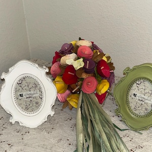 May include: A vibrant bouquet of fabric flowers in red, pink, yellow, and purple, with green detailing. The bouquet is next to two decorative oval picture frames, one white and one green, with the text "valerie bertinelli."