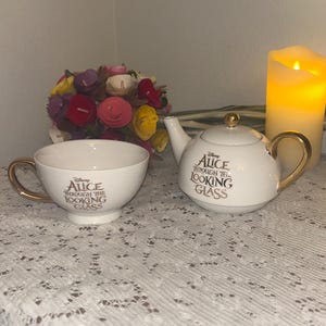 Puede incluir: Un juego de taza y tetera blancos con detalles dorados. El juego presenta la inscripción "Alice Through the Looking Glass" en letras doradas. Un ramo de flores coloridas y una vela encendida están en el fondo.