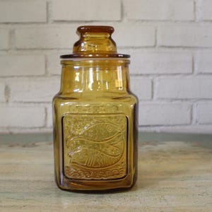 Puede incluir: Tarro de cristal color ámbar con tapa. El tarro tiene forma rectangular con un panel decorativo en relieve con un diseño de pez. El tarro está sobre una superficie de madera frente a una pared de ladrillos blancos.