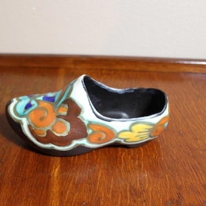 May include: A ceramic Dutch clog-shaped decorative piece. The shoe is black on the bottom and inside, with a white top and colorful floral designs in orange, brown, blue, and yellow. The piece is sitting on a wooden surface.