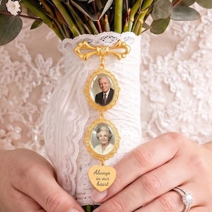 Può includere: Un ciondolo commemorativo dorato con due cornici ovali contenenti i ritratti di un uomo e una donna anziani, sospeso a una spilla a fiocco decorativa. Un ciondolo a forma di cuore inciso con Always in our heart è fissato a un manico di bouquet avvolto in pizzo bianco.