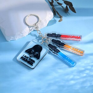 May include: A keychain featuring a black Darth Vader design with the text "I AM THEIR FATHER." Attached are three lightsaber-shaped keychains with the names "SOPHIA," "CAESAR," and "ANNA." The keychains are made of clear acrylic.