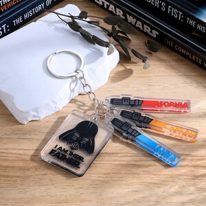 May include: A collection of Star Wars-themed keychains. One keychain features Darth Vader with the text "I AM THEIR FATHER." Other keychains are shaped like lightsabers, with names like "Sophia," "Caesar," and "Anna."