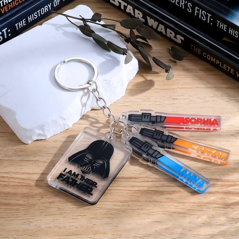 May include: A collection of Star Wars-themed keychains. One keychain features Darth Vader with the text "I AM THEIR FATHER." Other keychains are shaped like lightsabers, with names like "Sophia," "Caesar," and "Anna."