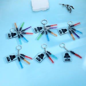 May include: Keychains featuring a black Darth Vader design with the text "I AM THE FATHER." Each keychain includes colorful lightsaber charms and name tags. The keychains are on a light blue surface.