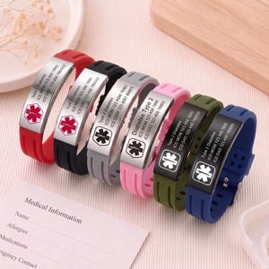 Könnte beinhalten: Ein Set aus sechs medizinischen Notfallarmbändern aus Silikon in Rot, Schwarz, Grau, Pink, Olivgrün und Blau. Jedes hat eine Metallplatte mit einem roten medizinischen Sternsymbol und Gravuren wie Type 2 Diabetes und Telefonnummern.