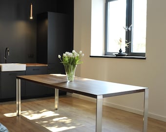 May include: A modern dining table with a dark wood top and stainless steel legs. The table is rectangular and sits on a light wood floor. A vase of white tulips sits on the table. The room has a minimalist design.