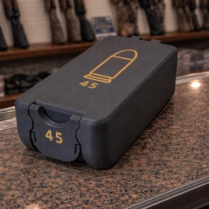 May include: A dark gray ammunition case with a gold bullet graphic and the number "45". The case is rectangular with a hinged lid and a latch. The number "45" is also displayed on the front of the case.