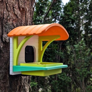 May include: A colorful bird feeder with an orange roof, green supports, and a white body is mounted on a tree. The feeder has a tray and a built-in camera. The background is blurred green foliage.