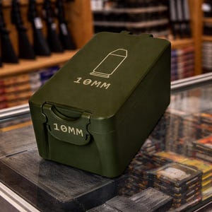 May include: An olive green ammunition box with the text "10MM" printed on the front and top. A white graphic of a bullet is on the top. The box is on a glass surface, with rifles and shelves in the background.