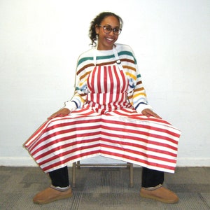 May include: A woman wearing a red and white striped apron over a rainbow striped sweater. She is sitting on a chair with her legs crossed and smiling at the camera.