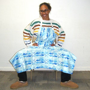 May include: A light blue apron with a seahorse pattern. The apron is worn over a striped sweater. The seahorse pattern is in shades of blue on a light blue background. The apron has a white neck strap.
