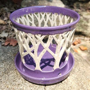 May include: A purple ceramic planter with a white tree-branch design cut out of the sides. The planter sits on a matching saucer. The rim of the planter and saucer are a solid purple color. The planter is designed for indoor or outdoor use.