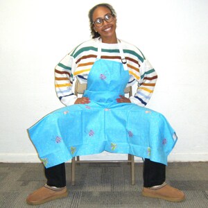 May include: A person wearing a blue apron with a butterfly pattern, a striped sweater, and glasses. The apron has a bib and two straps. The person is sitting on a chair with their legs crossed.