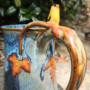 May include: A ceramic mug with a blue and cream glaze, featuring orange leaf accents. The handle is brown and the top of the handle has a yellow bird figurine. The mug is designed for beverages.