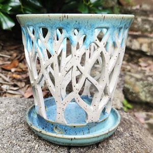 May include: A ceramic planter with a unique tree branch design. The planter is white with a blue glaze on the rim and base. The design features cut-out branches, and it includes a matching saucer.