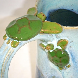 May include: A blue ceramic mug with a green turtle handle. The turtle is sculpted in a 3D design and has a glazed finish.