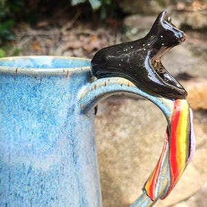 May include: A handmade ceramic mug with a blue glaze and a black cat figurine on the handle. The handle is decorated with colorful stripes. The mug is a unique piece of functional art.