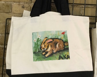 May include: White canvas tote bag with black handles and a black bottom. The bag features a painted image of a brown rabbit in a field of green grass and white flowers. The text "AKA" is printed in black below the rabbit.