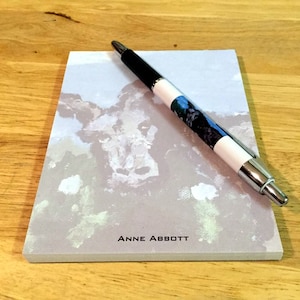 May include: A grey notepad with a watercolour painting of a cow's face on the front. The notepad has the text "ANNE ABBOTT" printed on the bottom. A black and silver pen is lying on top of the notepad.