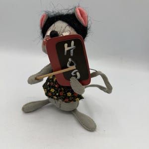 May include: A grey felt mouse doll with a black wig and pink ears, holding a small red chalkboard with the word "Hog" written in white. The mouse wears a floral print dress and has a wooden stick attached to the chalkboard.