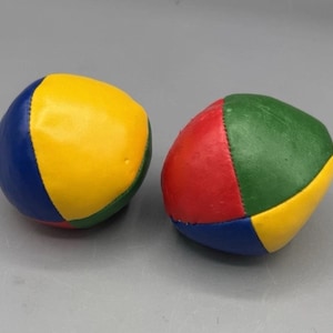 Puede incluir: Dos pelotas de malabares multicolores. Una pelota presenta paneles azules, amarillos y verdes. La otra pelota tiene paneles rojos, verdes y amarillos. Ambas están hechas de un material suave y cosido.