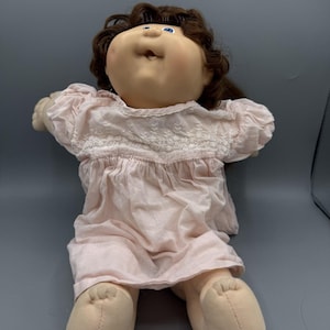 Puede incluir: Una muñeca vintage con una cara suave y redonda, ojos azules y cabello castaño rizado. La muñeca lleva un vestido rosa claro con detalles de encaje. Los brazos y las piernas de la muñeca son de tela beige suave. La muñeca está tumbada sobre una superficie gris.