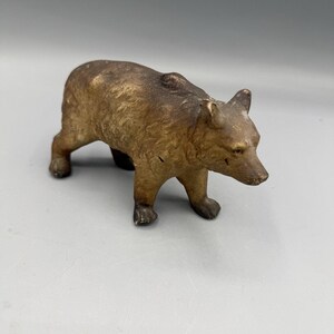 May include: A small, antique-style bear figurine in shades of brown. The bear is depicted in a walking pose, with its head slightly lowered. The figurine has a textured surface, giving it a vintage appearance. It is a decorative item.