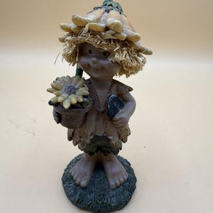 May include: A figurine of a small character wearing a hat made of yellow sunflower petals and straw-like fringe. The figure has brown skin, wears a brown vest and green shorts, and holds a small potted sunflower and a black hand trowel.