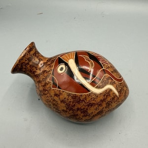 May include: A brown, fish-shaped ceramic vessel with a textured surface. The vessel features a detailed design of a fish with red, black, and white accents. The vessel has a small opening at one end and a rounded body.