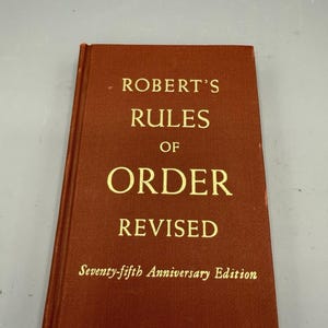 May include: A brown book titled "Robert's Rules of Order Revised" with gold lettering. The cover also includes "Seventy-fifth Anniversary Edition". The book is a guide to parliamentary procedure.