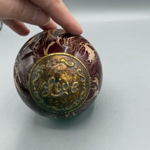 May include: A decorative, spherical object with a maroon and cream marbled pattern. A circular, antique gold-coloured metal plate is affixed to the sphere, with the word "Live" in an ornate script. The object is being held by a hand.