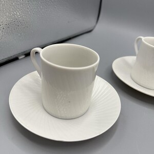 May include: Two white ceramic teacups and saucers. The cups are cylindrical with small handles. The saucers have a radial, textured pattern. The set is clean and minimalist, ideal for serving tea or coffee.
