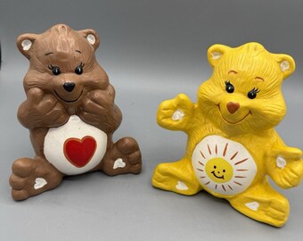 May include: Two ceramic Care Bear figurines. One is brown with a red heart on its belly. The other is yellow with a smiling sun on its belly. Both have black eyes and are sitting down.