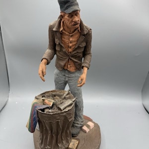 Puede incluir: Figura detallada de un anciano con ropa desgastada, de pie junto a un cubo de basura lleno de escombros. El hombre lleva una gorra oscura, una chaqueta marrón y pantalones grises. Un paño colorido cuelga del cubo de basura.