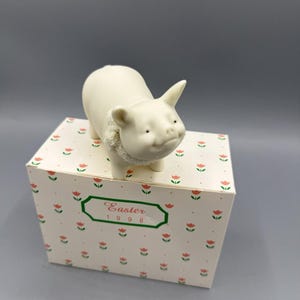 May include: A small, off-white ceramic pig figurine with a textured collar, standing on a decorative box. The box is white with a pattern of red tulips and the text "Easter 1998" in a green frame.
