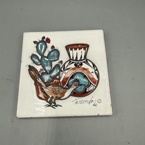 May include: Square ceramic tile with a Southwestern design. The tile features a blue cactus with red flowers, a brown roadrunner, and a decorative vase with black, red, and white patterns. The tile is signed "Teissedre 92".