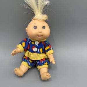 Könnte beinhalten: Eine Vintage-Cabbage-Patch-Kids-Puppe mit kahlem Kopf, blauen Augen und einem kleinen Lächeln. Die Puppe trägt ein blau-gelb gemustertes Outfit mit Rüschenkragen und passenden Shorts. Ihr blondes Haar ist zu einem Stachel nach oben gestylt.